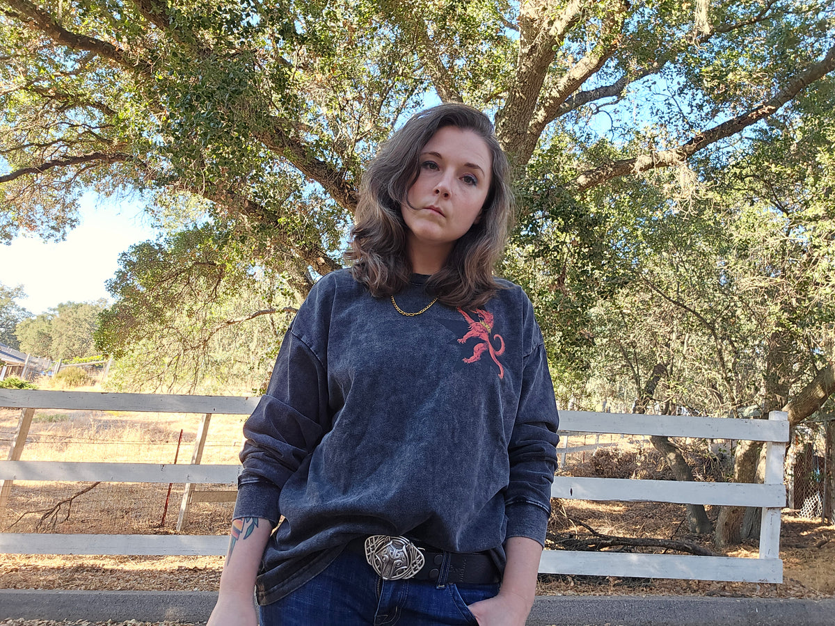 A woman wearing a dark washed blue sweatshirt with a red embroidered logo on the left chest, paired with blue jeans and a silver belt buckle, standing outdoors in front of a white fence and a large tree.