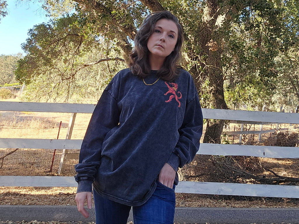 A woman wearing a dark navy blue long-sleeve sweatshirt with a red embroidered logo and blue jeans, standing outdoors in front of a white wooden fence and a large tree.