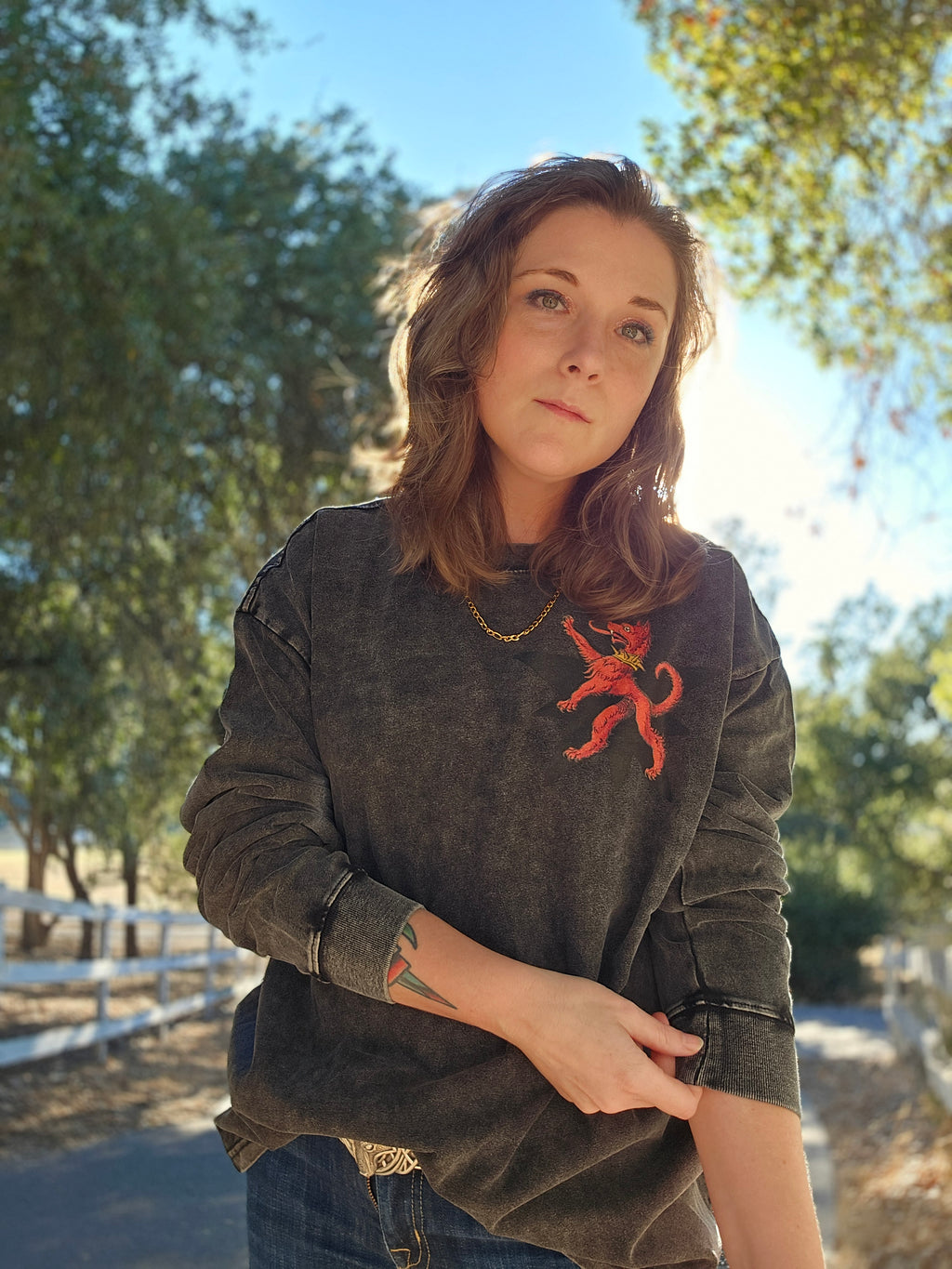 A woman wearing a dark gray washed crewneck sweatshirt with a red dragon graphic on the chest, standing outdoors on a sunny day with trees and a fence in the background.