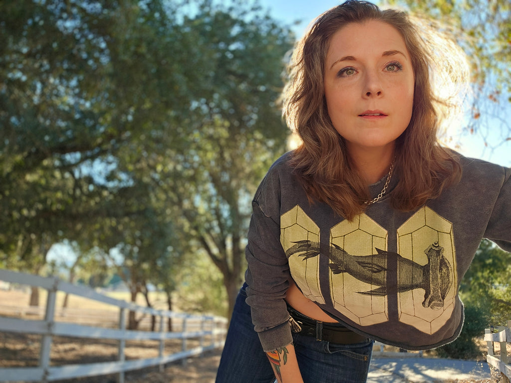 A woman wearing a dark gray graphic sweatshirt with a  green and black fish design, paired with blue jeans, photographed outdoors in front of trees and a white fence.