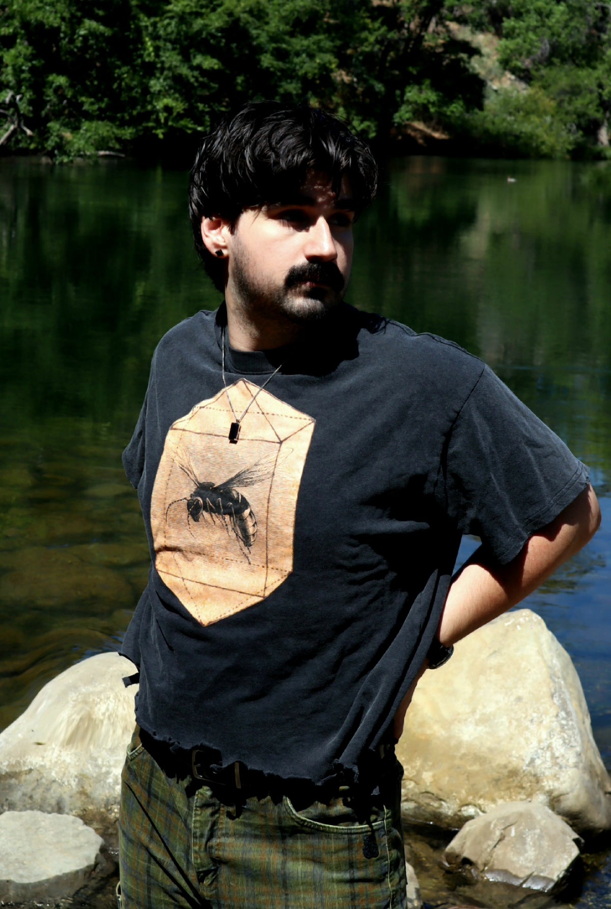 A man standing outdoors by a river wearing a dark charcoal t-shirt with a large orange wasp graphic featuring a black bee, paired with green plaid pants.