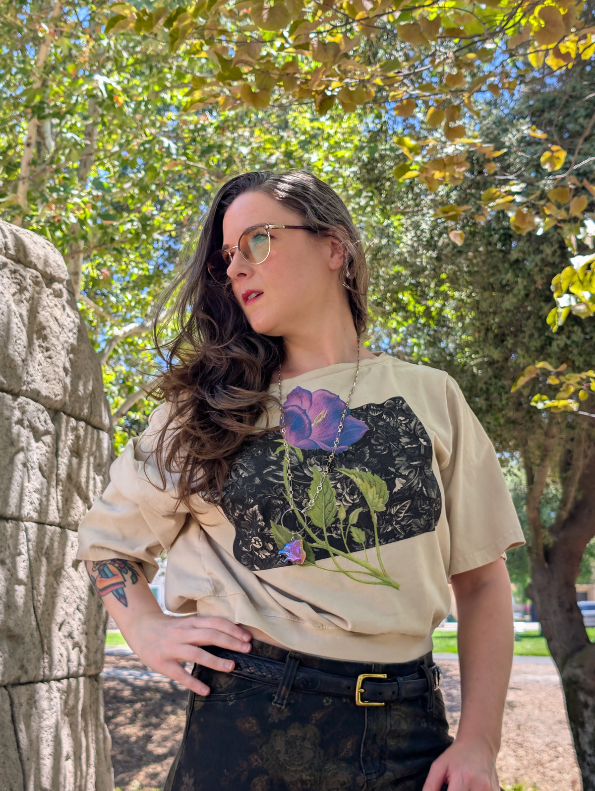 A woman wearing a beige graphic t-shirt with a purple flower design, styled with black jeans and sunglasses, standing outdoors in a park with trees and a stone wall in the background.