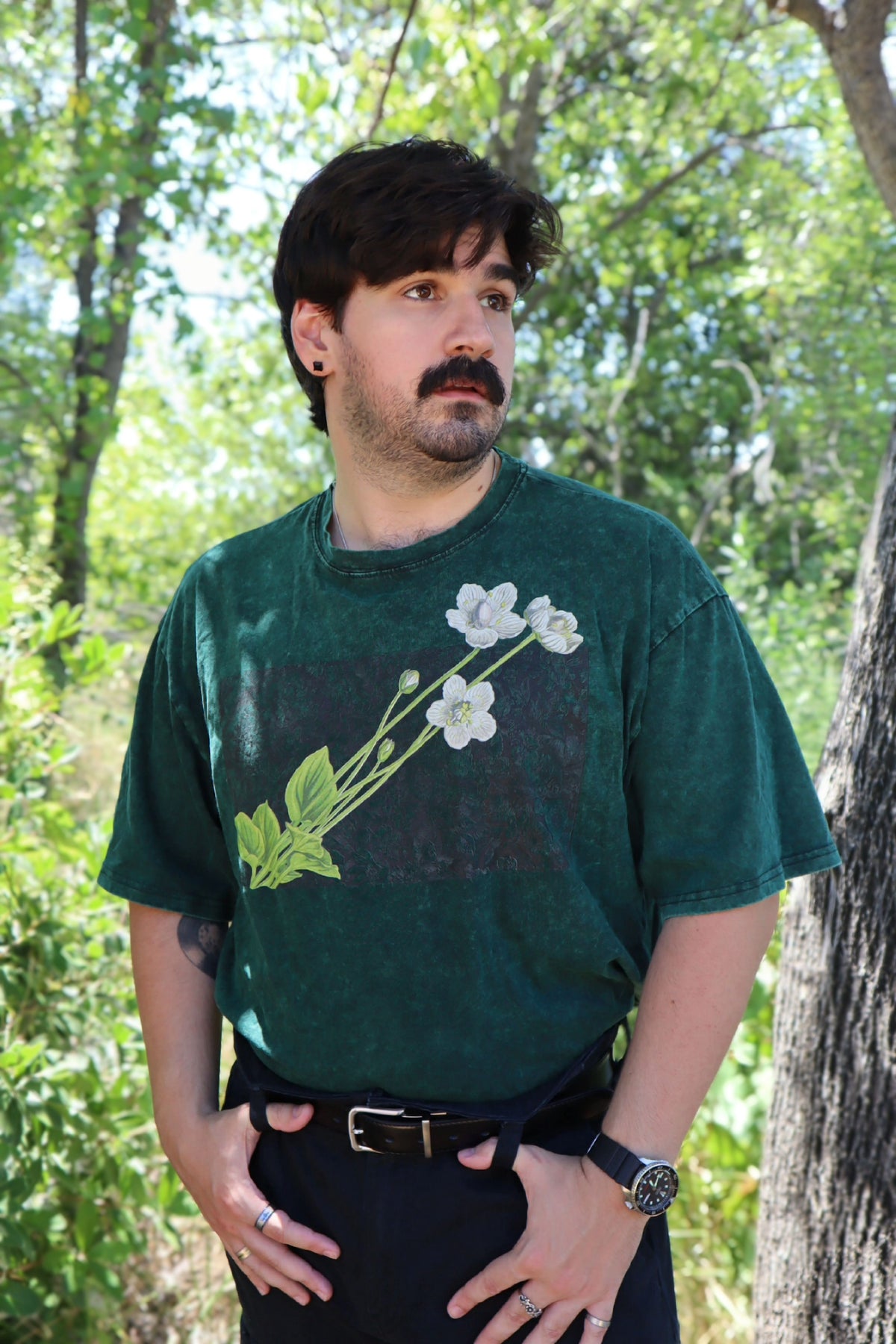 A man wearing a dark green vintage graphic t-shirt with a white flower and green leaf print, standing outdoors in a wooded area.