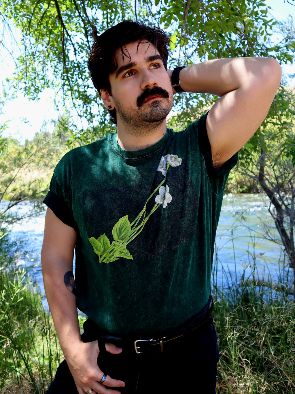 A man wearing a dark green graphic t-shirt with a white flower design, standing outdoors near a river with trees in the background.