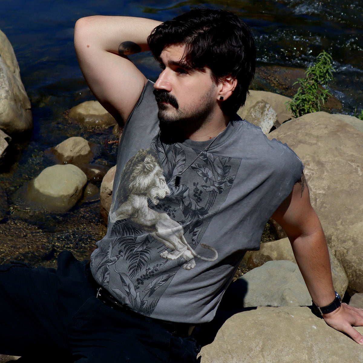 A man sitting on large rocks by a flowing stream, wearing a gray sleeveless graphic t-shirt with a lion print and dark pants.
