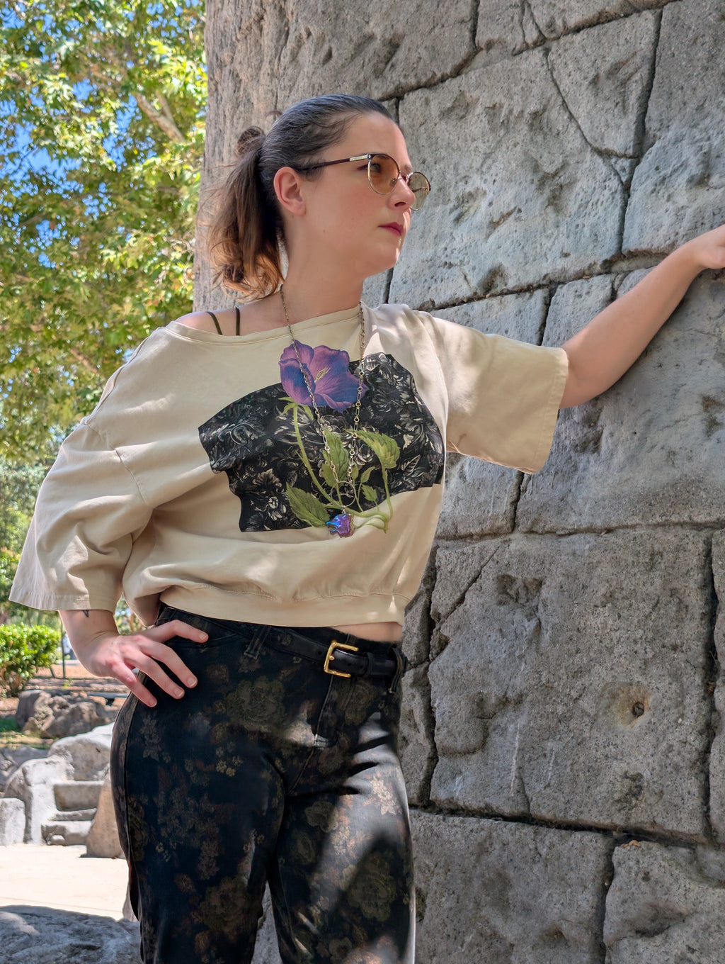 A woman wearing a beige oversized crop top with a purple and green floral graphic print, paired with dark patterned pants, posing outdoors against a stone wall.