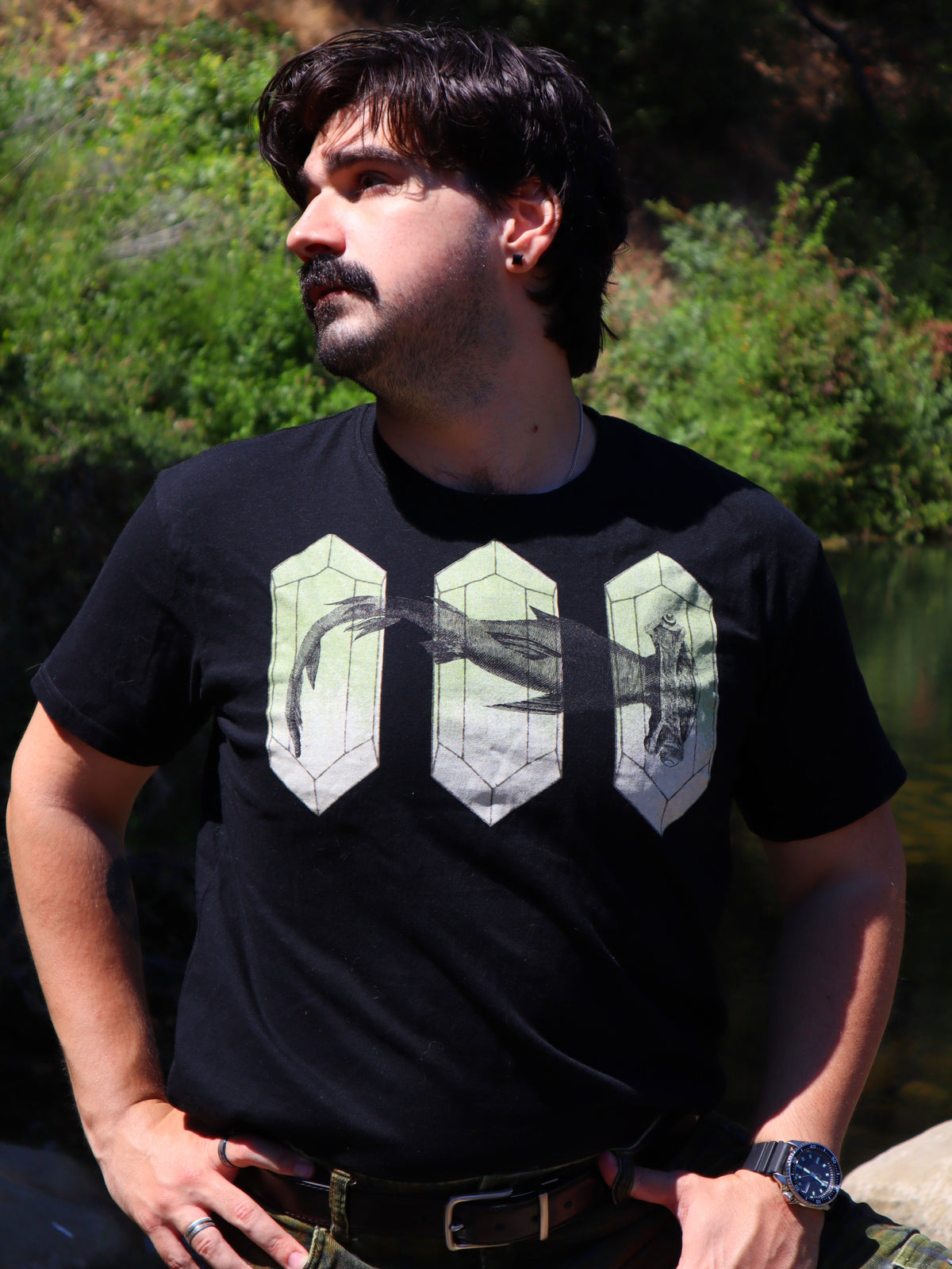 A man wearing a black t-shirt with a graphic print of three 3D geometric crystal structures containing a hammerhead shark, set against a natural outdoor background of green foliage and water.
