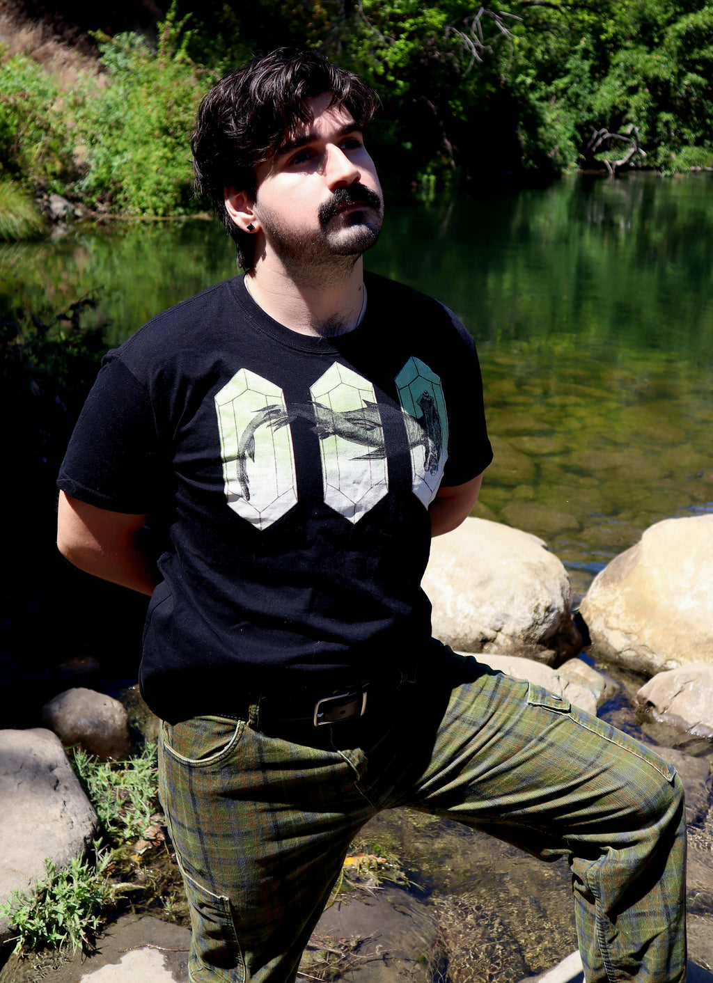 A man standing outdoors by a river wearing a black graphic t-shirt and green plaid pants.
