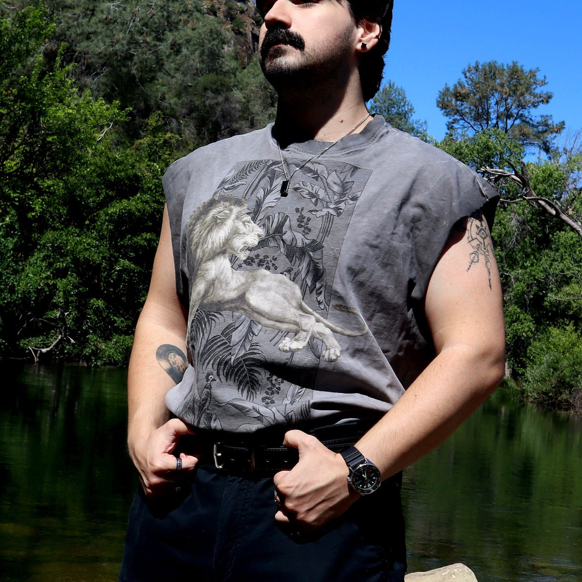 A man wearing a gray sleeveless graphic t-shirt with a detailed lion print, standing outdoors near a body of water with trees in the background.