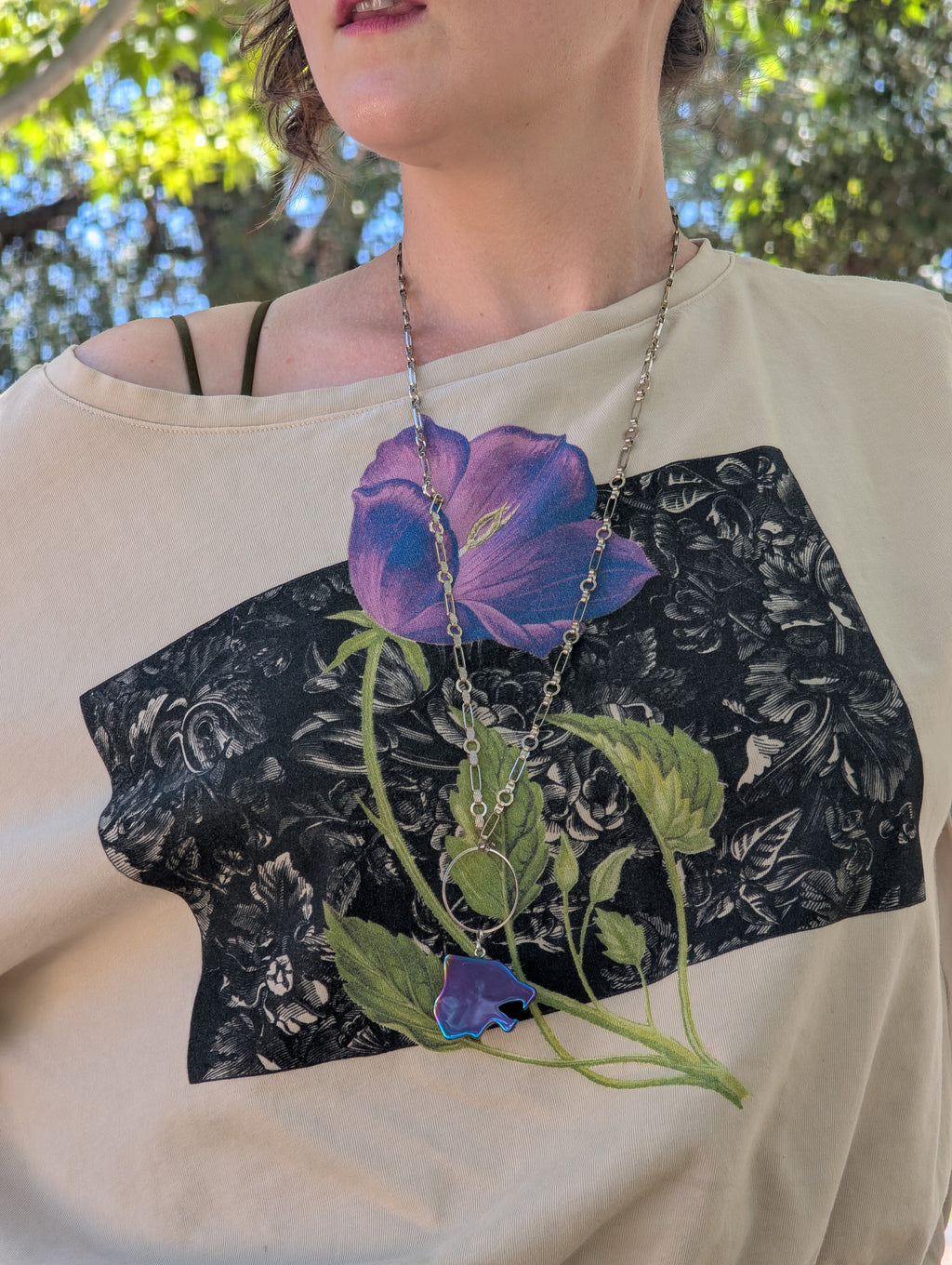 A close-up of a person wearing a cream-colored off-the-shoulder t-shirt with a graphic print of a purple and blue flower over a black floral lace pattern. The person is also wearing a silver chain necklace with a purple flower pendant.